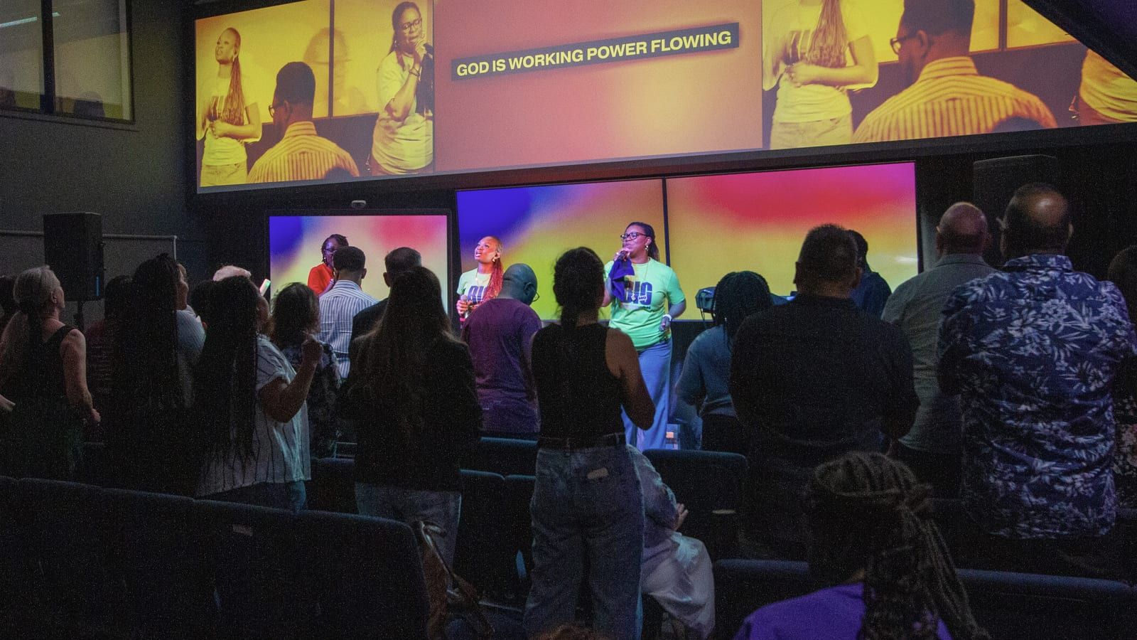 The room from the back during worship: the congregation standing, and the singers on the platform beneath the screens.