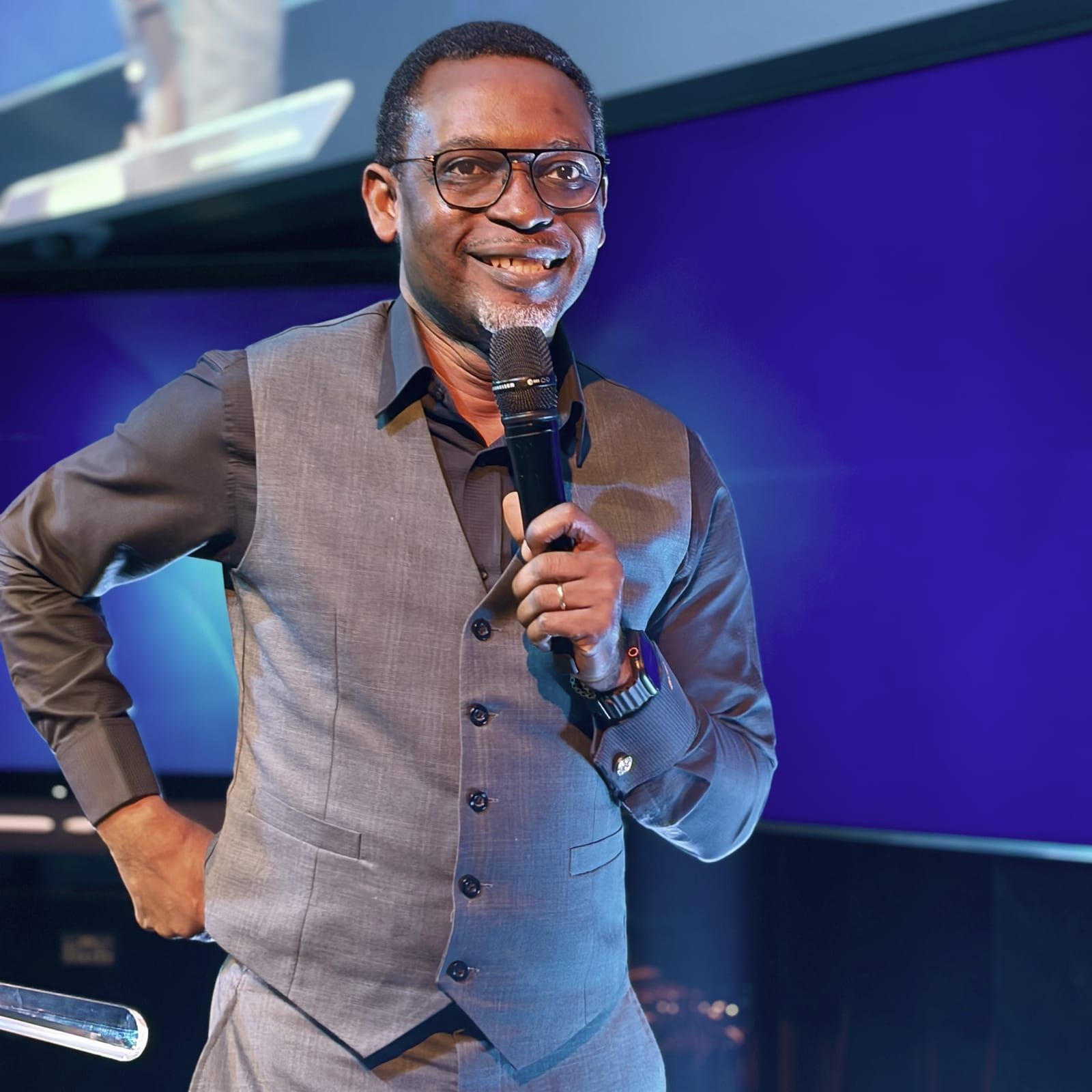 Dr Michaels Aibangbee, microphone in hand, smiling mid-sentence on the platform.