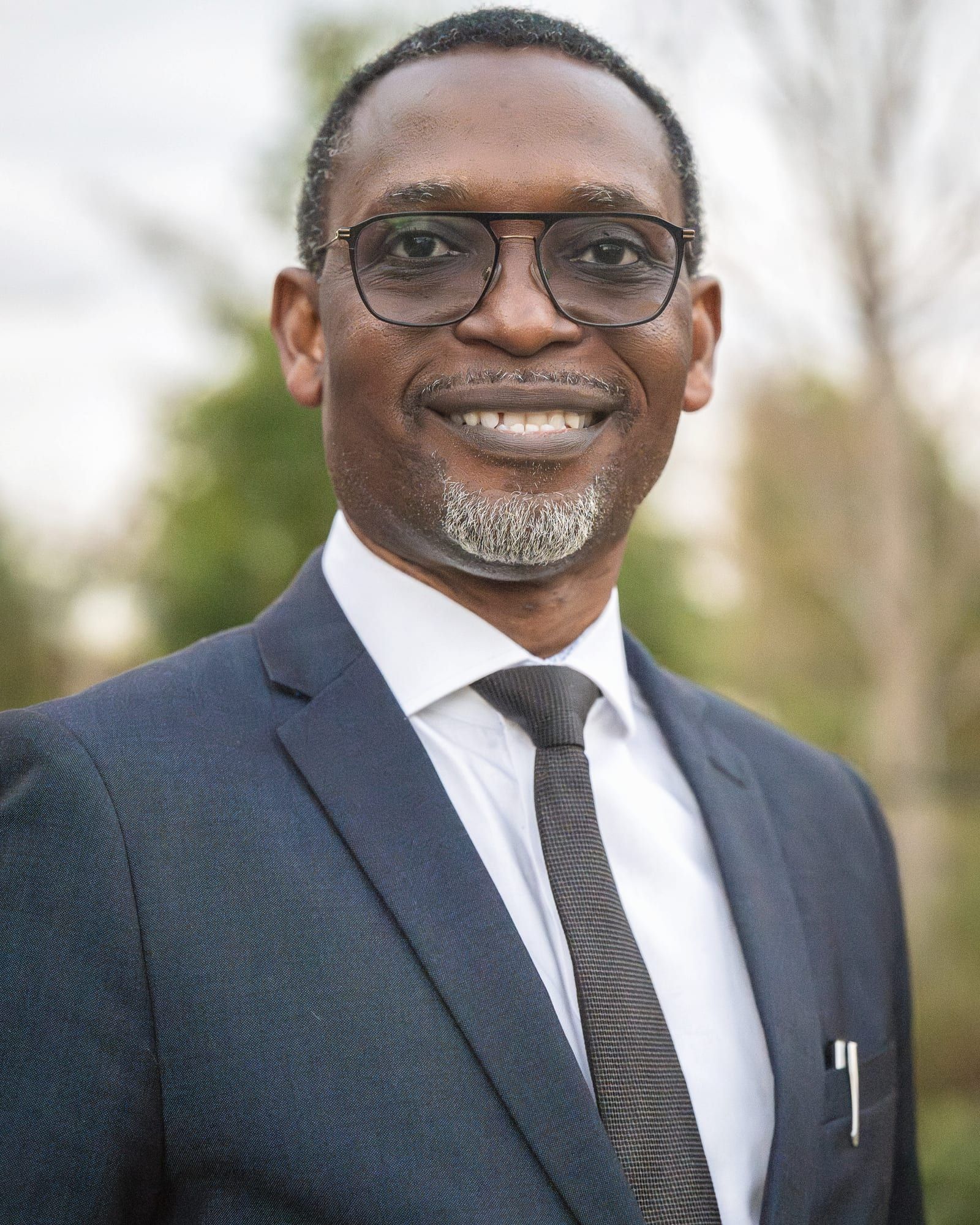Dr Michaels Aibangbee smiling, in a navy suit and tie, photographed outdoors among trees.